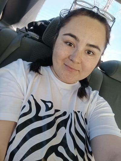 A young woman is pictured in a selfie taken inside a vehicle. She is wearing a white t-shirt with a black zebra print design. Her dark hair is styled in braids, and she has glasses perched on top of her head. The background shows the interior of the car, with grey upholstered seats and a glimpse of the sky visible through the window. The scene appears to be taken during the daytime, suggested by the daylight. There are no indicators to place this in Ust-Kishert, Russia.
