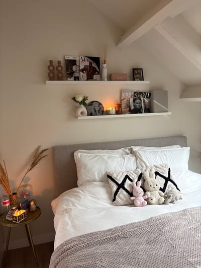 This image captures a cozy and neatly arranged bedroom interior, located in Rosmalen, Netherlands. The photo is oriented ninety degrees clockwise, depicting a wall-mounted shelf on the left side and a bed with a bedside table on the right.
The light-colored wall features a white wall shelf, adorned with various decorative and functional items. The upper tier holds a grey box, a framed photo, and several magazines, one prominently displaying "ESTAGGI." Below, the lower tier of the shelf showcases a white diffuser with reeds, a white vase, more magazines with visible faces, a burning candle, a round black-and-white print of a person's face, a white ceramic donut-shaped object, and a small arrangement of white roses and green foliage. Below the shelf, two wooden, circular decorative pieces hang on the wall. The wall continues into a sloped ceiling with visible white beams.
To the right, a bed features a grey upholstered headboard and white bedding, including a duvet and pillows. A textured white pillow with a bold black abstract 'X' pattern rests on the bed, accompanied by three plush toys: a grey animal, a white bear, and a pink bunny. A light grey, knitted throw blanket partially covers the bed.
In the bottom right corner, a small, round, gold-toned bedside table with slender legs holds a clear vase filled with dried pampas grass, a clear water bottle, a small house-shaped candle holder with a glowing light inside, and a small figurine next to what appears to be a small book or box.
The lighting, particularly from the burning candles, suggests an evening or low-light setting, contributing to a warm and inviting atmosphere. There are no people visible, and no specific activity is taking place, emphasizing the serene and styled domestic scene. The overall color palette is predominantly neutral, featuring white, various shades of grey, and natural wood tones, accented by hints of black and the soft pink of the bunny.