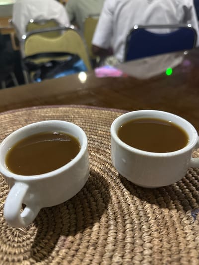 Two white demitasse cups filled with dark brown liquid, presumably coffee, are positioned on a woven straw mat on a wooden table. The cups are arranged diagonally, with the one on the left closer to the viewer and slightly to the left, and the one on the right further away and to the right. The background is blurred, showing an indoor setting with people sitting in chairs. The lighting suggests an artificial indoor environment, possibly a cafe or restaurant. The scene is devoid of any explicit identifiers placing it in Dandagoro, Nigeria, or any other specific location.