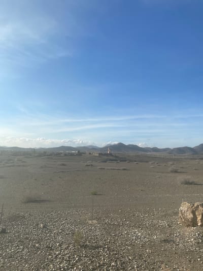 The image displays a vast, arid mountainous landscape under a bright, partly cloudy sky. The photograph, presented rotated 90 degrees clockwise, depicts a wide expanse of stony ground in the foreground and mid-ground, giving way to a range of rugged mountains in the distance.
The upper half of the correctly oriented image features a clear blue sky, interspersed with long, wispy white clouds stretching horizontally across the frame. Below the sky, a continuous chain of dark, rolling mountains forms the distant backdrop, appearing somewhat hazy. Within the mountainous terrain, a small cluster of buildings is discernible, with one structure prominently featuring a minaret, suggesting a mosque.
The foreground and mid-ground consist of barren, rocky, and gravelly terrain, predominantly in shades of grey and brown. Sparse, low-lying desert vegetation, appearing as small shrubs or bushes, is scattered across this stony expanse. A notable, lighter-colored rock outcrop is visible on the right side of the foreground.
The scene conveys a sense of tranquility and vastness on a clear, sunny day, likely morning or late afternoon, given the soft, ambient lighting. There is no visible human activity or text. The landscape is characteristic of the Jbilate region in Morocco, confirming its arid and mountainous nature.