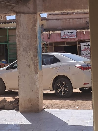 A white sedan is parked in front of a building located at 18.544, 31.848 in Karima, Sudan. The car is facing the right. The car's front left tire is visible, and the back right tire is fully visible. The right side of the car is visible, with the back door and the back side of the car visible. The rear passenger door is visible. The car is parked on a dirt road with a concrete sidewalk to the right. There is a large, weathered concrete pillar to the right of the car. The pillar is partially obstructing the view of the car. The car is in the shade, and the ground is covered with dirt and a few bricks. The building is partially visible and appears to be a shop. The shop has a red roof with a white sign above the door. The sign is in Arabic. There is a small, red sign with Arabic writing on it near the back of the car. The sign is partially obscured by the pillar. The sky is clear and blue, and there are some buildings in the background.  There are no people present.  The location is the outskirts of Karima, Sudan.
