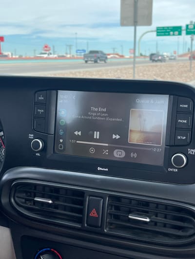 The image captures the interior of a vehicle, specifically its dashboard and infotainment system, with a blurred view of an urban roadway through the windshield. The infotainment screen, displaying a time of 10:47 and a 5G cellular signal, shows a music application playing "The End" by Kings of Leon from the album "Come Around Sundown (Expanded)." The track is 1 minute and 57 seconds into its playback, with 2 minutes and 27 seconds remaining. Physical controls surrounding the screen include buttons for "SEEK," "TRACK," "HOME," "PHONE," "SETUP," "ENTER," and a "PWR PUSH VOL" (volume) knob. Below the screen are horizontal air vents and a red triangular hazard light button. The exterior environment, observed through the windshield, depicts a multi-lane highway or major road under an overcast sky. Several vehicles, including a grey pickup truck, are visible on the road. Green overhead road signs are discernible, with one clearly indicating "I-10" and another pointing towards "Lee Trevino Dr" and "Pellicano Dr." This identifies the location as the vicinity of Interstate 10 near the Lee Trevino Drive and Pellicano Drive interchange in El Paso, Texas, United States. No individuals are visible in the image.