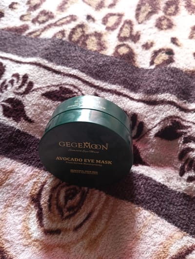 The image shows a dark green jar of GegeMoon Smooth Eye Mask with Avocado Eye Mask and Fruit Serum Moisturizing text printed on it. The jar is placed on a patterned fabric with leopard print details. The scene is indoors and the lighting suggests it is daytime. There are no people or animals visible in the image. The location is not specified as Monastirische, Russia; this information is not present in the image.