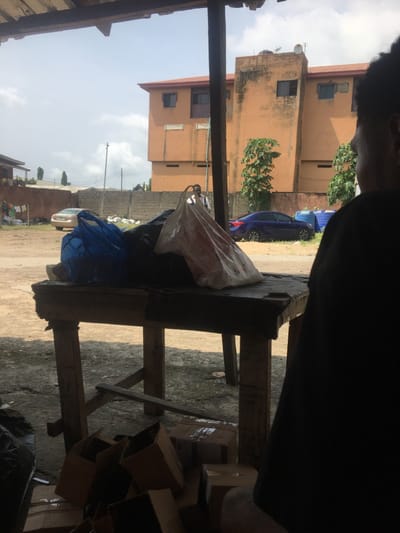 The scene depicts an outdoor or semi-outdoor area in Awjaw, Nigeria, under a wooden shelter, viewing an unpaved open space and residential structures under a partially cloudy sky. In the immediate foreground, to the right, the back and right shoulder of an individual wearing a dark garment are visible, facing left. Central to the foreground is a weathered wooden table laden with multiple plastic bags: one blue, one black, and one white with red markings. Beneath the table, a pile of brown cardboard boxes rests on the ground.
Beyond the immediate foreground, an unpaved dirt path or open lot is visible. On the left side of this path, a light-colored sedan is parked near a low concrete block wall, behind which clothes hang on a line. Towards the right, a dark blue sedan is parked. Between the two vehicles, a male figure in a light shirt and dark trousers stands facing generally right.
The background is dominated by a large, multi-story building with an orange-ochre facade and a reddish-brown roof, featuring multiple windows. The building shows signs of weathering and discoloration. A satellite dish is visible on its roof. Trees with green foliage are present near the building. Overhead, parts of the wooden beam and slat structure of the shelter are visible, indicating the vantage point.