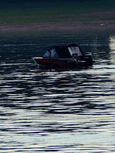A black and red motorboat with a black canopy is moving on dark water, leaving a wake. The boat has a Suzuki outboard motor. The background shows a shoreline with green vegetation. The overall scene is likely a natural body of water, possibly a lake or river, and the image does not provide any specific information about the city of Erkalovo, Russia.