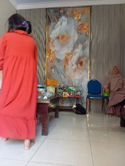 The scene depicts an indoor room located in the city of Pasuruan, Indonesia. In the foreground, a barefoot individual with dark hair secured by a purple clip, wearing a long, reddish-orange dress with a ruffled collar, is captured from behind, facing away from the viewer and appearing to move towards the left. To the right, a second individual is seated on a dark wooden chair, positioned next to a blue metal chair. This person wears a light brown hijab and matching attire, with a mauve-colored fabric draped over their lap, and faces slightly left. Between the individuals, a dark brown wooden rectangular table holds a large yellow lidded container, a clear glass jar filled with small colorful items, and other assorted containers. To the right, a smaller table displays several snack boxes, and a black bag rests on the tiled floor beneath it. The background wall features a large, vertically oriented framed artwork depicting prominent white flowers with golden details against a grey, textured background. The surrounding walls exhibit a vertical striped texture, and the floor consists of light-colored, glossy tiles. A white ceiling with decorative molding is visible.
