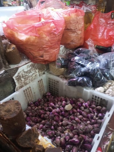A low-angle shot displays a variety of food items in a market setting. In the foreground, a white plastic crate is filled with red onions. Large brown blocks are beside the crate. In the background, translucent red plastic bags hold assorted goods. Small black bags containing dates and garlic bulbs are visible between the red bags and the crate. A bottle of oil is seen amongst the goods. A tiled surface is visible behind the food.