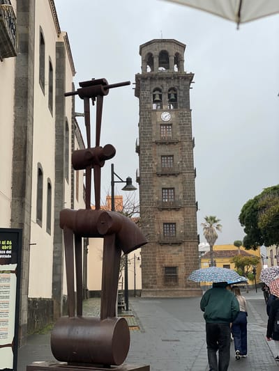 The image captures an overcast street scene in San Cristóbal de La Laguna, Spain, characterized by a historic bell tower, a contemporary sculpture, and pedestrians navigating wet pavement with umbrellas.
Dominating the background is a sturdy bell tower constructed from dark, possibly volcanic, stone. It features a multi-tiered design with arched openings for bells at the top, and a prominent white clock face with black Roman numerals below. Rectangular windows with dark frames are symmetrically placed along its mid-section.
In the foreground, an imposing abstract sculpture crafted from dark, rusted metal, likely iron or steel, stands. It consists of thick, angular, and cylindrical components, giving it an industrial or abstract figurative appearance, with hammer-like forms and connecting rods.
Several individuals are visible, walking away from the camera on the visibly damp, paved street. They are holding umbrellas, indicating rain or recent precipitation. One person wears a dark green jacket and carries a light blue umbrella adorned with white and pink patterns. Another person uses a patterned, possibly multi-colored, umbrella.
The surrounding architecture includes buildings with light-colored, cream-toned facades and dark stone bases or framing. Distant greenery, including palm trees and other trees, adds to the urban landscape. A white awning or canopy is partially visible on the far left. On a dark poster board in the lower right, partially readable text includes "al 26 abril," "Plaza del Adelantado," "TITIRITANDO ARTEFACTOR," "PEPE LUNA," and "La Concepción," suggesting details for local cultural events or a festival.