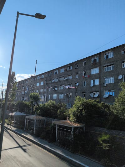 The image depicts an apartment building with a gray, weathered facade, its windows adorned with various shades of blue, white, and some with laundry hanging out to dry. The building's architecture suggests an older, possibly Soviet-era construction. On the right side of the frame, dense green trees partially obscure the building, casting shadows. A paved road runs alongside the building, with a sidewalk bordered by a low, grey wall. Several wire cages or enclosures are visible behind the wall, some covered with a light-colored material. A street lamp with a distinctive curved arm extends from the left side of the image into the clear blue sky. The overall scene suggests a residential area on a sunny day, likely in the afternoon given the shadows. The presence of satellite dishes on the building hints at modern amenities in an older setting. The location context provided is Şəki, Azerbaijan.