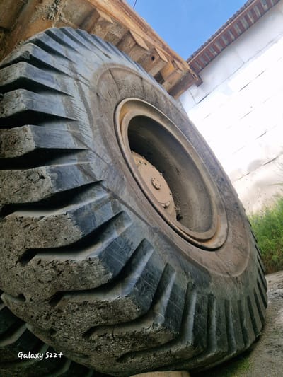 This is a low-angle, close-up photograph of a massive, heavily worn tire of what appears to be heavy industrial machinery, possibly a dump truck, situated outdoors. The tire is black, deeply grooved, and covered in dust and dirt, indicating significant use. The rim and central hub assembly are visible, with a rusty metal cover. In the background, there is a weathered, rusty metal structure with exposed beams and supports, likely part of the same vehicle or a nearby industrial building. To the right, a white wall or building facade is visible, with a corrugated metal roofline above it. Patches of green grass and foliage can be seen behind the tire. The sky above is a clear blue. The photograph is taken from a low perspective, emphasizing the immense scale of the tire. The text "Galaxy S22+" is imprinted in the lower left corner, indicating it is a photograph taken with a Samsung Galaxy S22+ phone. No people are visible in the image. The location is identified as Zheleznogorsk-Ilimsky, Russia.