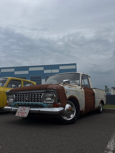 In Uzda, Belarus, a heavily rusted white pickup truck with a white cab and a light blue grille is parked on an asphalt surface. The truck has a dark metal fender and a chrome bumper with a license plate that reads "CH 6780". A yellow sedan is parked to the left of the truck. In the background, a building with blue and white checkered patterns is visible. The sky is overcast with gray clouds.