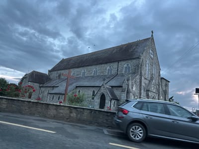 A grey station wagon drives past a stone church under a cloudy sky in an unknown town in Ireland. The church has a tall, pointed roof and arched windows. A large wooden cross stands in front of the church, and red roses bloom along a stone wall. The road in front of the car has a yellow line. There are birds flying in the sky above the church.