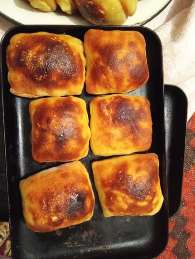 The image shows a black baking tray filled with six golden-brown, square-shaped pastries. The pastries have a slightly shiny surface with some areas of darker browning, indicating they are freshly baked. Some of the pastries are sprinkled with sesame seeds, which are visible as small, light-colored dots. The baking tray itself appears to be well-used, with some scuff marks and a slightly worn appearance.

In the background, out of focus, there are more pastries on a white plate, appearing lighter in color and possibly different in shape. A white textured cloth is also visible in the background, along with a patterned rug with red and brown tones, suggesting a home setting. The lighting suggests it could be daytime, likely indoors, as the lighting is somewhat diffused. There is no visible text in the image. The scene depicts a collection of baked goods, possibly a meal or a treat, prepared in a domestic environment in Жошук, Kyrgyzstan.