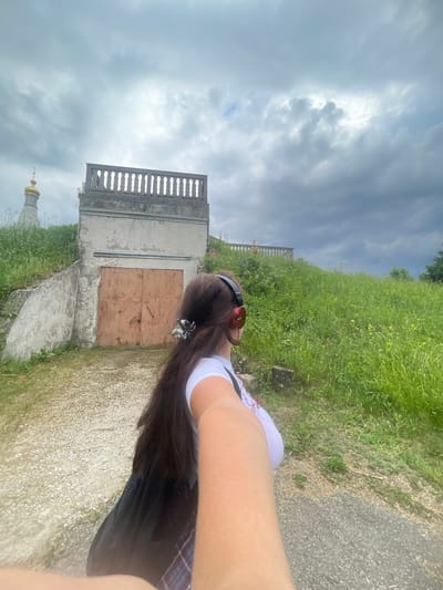 An overcast day unfolds in Frontoviki, Russia, with a woman in a white t-shirt and red headphones, holding a phone for a selfie. She stands on a gravel path, her long brown hair adorned with a metallic clip, obscuring her face as she turns away from the camera. A black bag hangs from her shoulder. Behind her, a weathered concrete structure with large, rusty metal doors dominates the foreground. To the left, a sloped concrete wall covered in graffiti leads up to the structure. Further in the background, a green hill covered in lush grass and wildflowers rises, crowned by a stone balustrade. A distant golden-domed structure, possibly a church, is visible atop the hill to the left. The sky is filled with dramatic, dark grey clouds, hinting at impending rain.