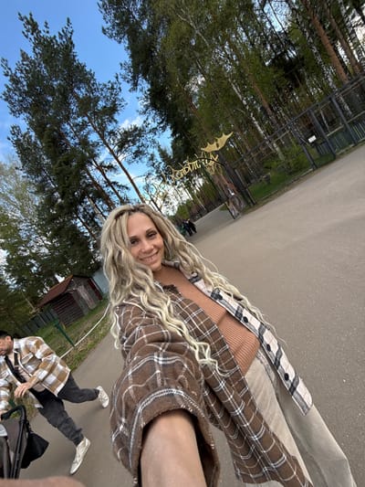 A woman with blonde dreadlocks smiles in a selfie taken outdoors in a park. She wears a tan top and a layered plaid shirt. Behind her, a path leads away, lined by tall trees and greenery. A man is visible pushing a stroller, wearing a plaid shirt and dark pants. A decorative sign is partially visible in the background, partially obscured by trees, indicating the location is likely in Yaroslavl', Russia. The sky is blue with some clouds.