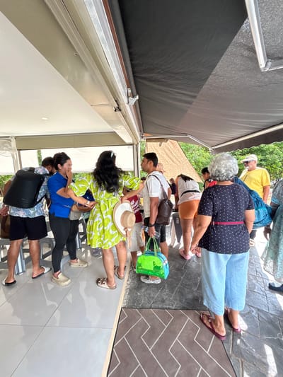 A group of people are gathered outdoors near a building, possibly a restaurant or cafe, in Ratsada, Thailand. The scene is partially covered by a dark awning. In the foreground, a woman in a lime green dress with white floral patterns is holding a straw hat. Next to her, a woman in a blue shirt and black leggings is speaking, with her hand near her mouth. A man wearing a black backpack and a floral shirt stands to their left. Further back, several other individuals are visible, some holding bags and others standing and observing. The ground is tiled with a pattern of light and dark grey squares. Trees and vegetation are visible in the background, suggesting a tropical environment. The overall impression is a casual gathering of people outdoors.