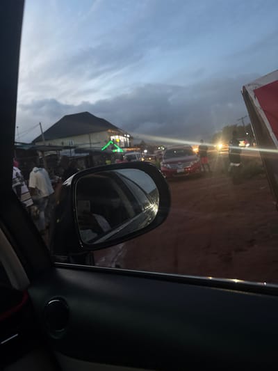 The image depicts a twilight scene in Ogheghe, Nigeria, viewed from inside a vehicle. The foreground shows the dark interior of a car door panel on the lower left, with a black side mirror occupying the central-left. The mirror reflects a portion of the vehicle's interior and a person's hand holding a white smartphone, positioned as if taking the photograph.
The scene outside the vehicle is an active, unpaved street or market area under an overcast, blue-grey sky. In the midground, a two-story building with a dark, pitched roof and light-colored walls stands prominently on the left. Below this building and extending to the left, numerous informal market stalls or structures are visible, with multiple individuals, including one wearing a white shirt, engaged in activity. Green neon-like lighting is visible on the building or an adjacent structure, along with other scattered artificial lights.
Further to the right, a red SUV is parked or moving slowly on the reddish-brown, unpaved ground, which shows signs of moisture or puddles. Several individuals are standing near this vehicle. In the background, vehicular headlights are visible further down the road, indicating traffic. The overall environment is a bustling commercial street at dusk.