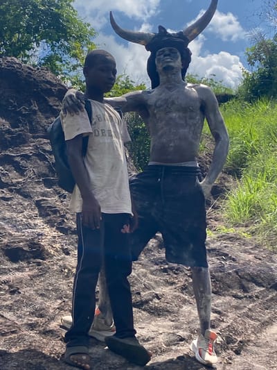 A man with dark skin stands in front of a statue of a man with horns, in Afikpo, Nigeria. The statue is of a muscular man with two large horns protruding from his head. The statue has on shorts and shoes. The man in front of the statue is wearing a white T-shirt, dark pants, and sandals. He has his arms around the statue. In the background, there is lush vegetation and a sky with clouds. The ground and area around them appears to be rocky terrain.