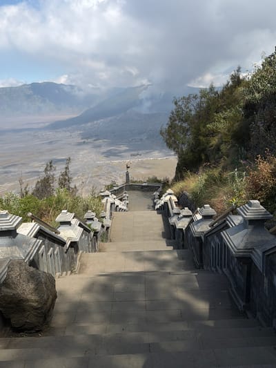 An upward-angled, full shot captures a long stone staircase leading down from a viewpoint overlooking a volcanic landscape in Cemorolawang, Indonesia. The stairs are bordered by intricately carved grey stone balustrades topped with ornate finials. To the right of the stairs, a steep, overgrown hillside is covered in dense green foliage, dry grasses, and some flowering plants. In the foreground, a large, dark grey boulder sits to the left of the staircase. Beyond the staircase and hillside, a vast, sandy plain stretches out, dotted with small white structures that appear to be tents or shelters. In the distance, layered mountains rise, shrouded in mist and clouds. A plume of smoke or steam drifts from one of the mountains, indicating volcanic activity. The sky is partly cloudy with patches of blue.