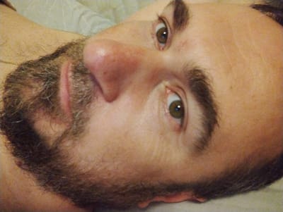 The image is a close-up, low-angle, indoor portrait of a fair-skinned man with brown hair and light brown eyes. The man is lying down, and the camera is positioned above and to his right. He has a light beard and mustache. His face is turned towards the camera, and his eyes are open. The background appears to be a light-colored, textured surface, possibly bedding. The lighting is dim and appears to be from a single source, casting shadows on the man's face. There is no visible indication of the location being in the city of Konstantinovo, Russia.