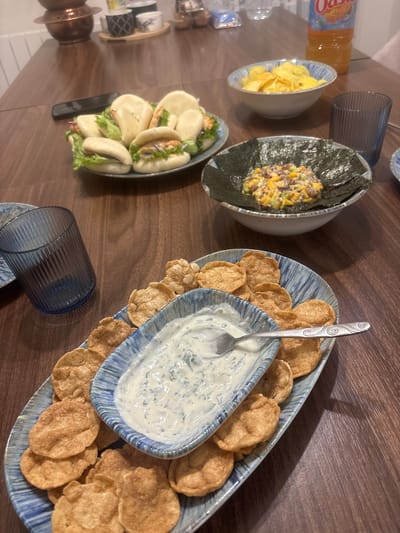 The scene depicts a dining table laden with various food items in a residential setting within Carrières-sous-Poissy, France. The table is constructed of dark, polished wood. No individuals are present in the image.
In the foreground, a long, narrow blue-and-white patterned ceramic platter is positioned, holding an array of golden-brown, circular fried crackers. Centered on this platter is a smaller, matching blue-and-white patterned ceramic bowl containing a white, creamy dip flecked with green herbs, into which a silver spoon with an ornate handle is resting. To the left of this platter, a dark blue glass with vertical ribbing is visible.
Towards the center of the table, a white ceramic bowl with a blue patterned rim holds a colorful mixture of diced ingredients, including yellow (likely mango), green (likely avocado), and reddish-purple (likely red onion). This mixture is encircled by several sheets of dark green nori seaweed.
Further back on the left side, a blue-and-white patterned ceramic plate presents approximately six white, steamed bao buns. These buns are split and filled with green lettuce and a light-colored protein, possibly garnished with thin slices of red onion. A black smartphone is partially visible on the table surface, positioned near the plate of buns.
On the upper right side of the table, a white ceramic bowl with a blue patterned rim is filled with yellow potato chips. Behind this bowl, an orange plastic bottle of "Oasis" brand juice is visible. A second dark blue glass with vertical ribbing is positioned to the right of the chips.
The background, slightly out of focus, reveals a light-colored wall and a shelf or surface displaying a copper vessel and several small decorative ceramic cups in varying patterns. A clear glass bottle of water is also discernible in the background.