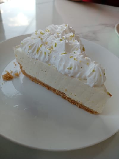 A close-up shot of a slice of key lime pie on a white plate. The pie has a graham cracker crust, a light yellow filling, and a whipped cream topping garnished with lime zest. The plate rests on a light-colored surface. The location is not discernible from the image. No people are visible.