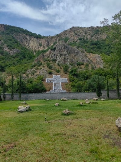 This media file captures a wide outdoor scene featuring a large cross monument set against a mountainous backdrop in Karnalovo, Bulgaria.
The primary subject is a monumental cross, constructed from grey stone or concrete, prominently displayed on the lower slope of a large, rugged mountain. The cross features a wide, stepped base and is composed of smaller, rectangular stone blocks, creating a textured surface. It stands out against the darker green foliage and rocky terrain of the hillside directly behind it.
The mountain itself dominates the upper two-thirds of the image, showcasing steep, rocky faces interspersed with dense patches of green deciduous and evergreen trees. The terrain is rough and natural, with visible rock formations and sparse vegetation higher up.
In the foreground, a vibrant green grassy field stretches across the bottom portion of the image. Several scattered, light-colored rocks of various sizes are visible on the grass. A low, grey stone wall or fence runs horizontally across the middle ground, separating the grassy field from the base of the mountain and the cross. Along this wall, several tall, slender evergreen trees (possibly cypress trees) stand at regular intervals.
The sky above the mountain is partly cloudy, with streaks of white and grey clouds against a light blue background, indicating daytime. No people or specific activities are visible in the image. There is no visible text or signage. The overall impression is one of a serene and significant religious or memorial site nestled within a natural landscape.