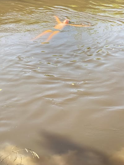 A person is floating on their back in murky, brown water. The person's arms are outstretched, and their legs are extended. The water has ripples and some floating debris, possibly leaves. The scene appears to be outdoors, with sunlight reflecting off the water's surface. The location is identified as Чэрвеньскі сельскі Савет, Belarus.