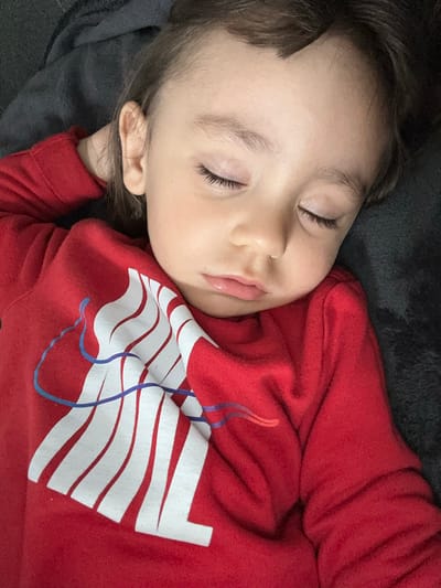 A close-up, eye-level shot captures a young child peacefully asleep on a dark, soft fabric, likely a blanket or pillow, in Fulda, Germany. The child's face is partially visible, turned slightly, showing closed eyes with long, dark eyelashes, a small nose, and slightly parted lips, indicating deep sleep. Their expression is serene and calm. Dark brown hair frames their face, resting against the dark background.
The child is wearing a bright red long-sleeved shirt or sweatshirt. The front of the shirt features a large, stylized graphic composed of white horizontal wavy stripes or a deconstructed flag-like pattern. Overlaid on this white design are blue and purple wavy lines, adding a dynamic accent. The background behind the child is a uniform dark grey or black, textured fabric, providing a soft contrast to the child's fair skin and red clothing. The lighting is soft and diffused, characteristic of an indoor setting during a quiet moment, such as a nap or night sleep. No explicit text is visible on the garment, only the abstract graphic. The overall scene conveys a sense of tranquility and innocence.
