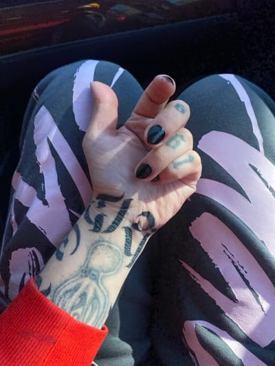 The image captures a close-up, downward-angled view of a person's left hand resting on their lap, located in Kruče - Kërruçi, Montenegro. The hand is light-skinned, with fingernails painted black. Tattoos are present on the fingers, displaying the characters "G", "1", and "B" (or "13"). The wrist and forearm are extensively tattooed with a faded grey octopus-like figure and intricate, dark, abstract or calligraphic patterns. A red, ribbed long-sleeved garment is visible on the left, covering the upper forearm. The lap is covered by dark grey or black trousers or leggings featuring large, abstract light pink or lavender designs. The upper background consists of a dark, reflective surface, likely the interior of a vehicle, showing indistinct reflections including a yellowish-orange area. Natural light illuminates the scene.