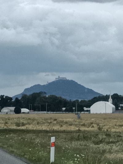 The image depicts a wide shot of a rural landscape under a cloudy sky. In the background, a prominent hill or mountain rises, crowned by a distant, somewhat indistinct structure that appears to be a castle or fortress. The middle ground features a line of dark green trees, behind which are several white structures, including a large tent-like building with a steeple and other smaller, translucent tent-like structures, possibly temporary event or storage facilities. The foreground consists of a field of dry, yellowish grass with scattered white wildflowers. A road is visible on the left edge of the frame, and a white road marker with a red reflective stripe is positioned in the grass. The overall impression is one of a vast, open space with a historical landmark in the distance. This scene is located in the vicinity of Bělá pod Bezdězem, Czechia, as indicated by the presence of the Bezděz Castle on the prominent hill.
