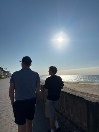 Two people stand on a brick path next to a beach. The person closest to the camera wears a blue polo shirt, dark shorts, and a baseball cap. The other person wears a black shirt and light-colored shorts. They both appear to be looking out at the water. The sun shines brightly in the sky, creating a glare. A concrete barrier separates the path from the sandy beach. Buildings are visible in the distance.