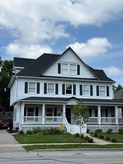 This is a full, eye-level, exterior shot of a white, two-story house with a black roof, black shutters, and a wrap-around porch. The house is located at 533 in Naperville, United States of America. A walkway leads from the sidewalk to the porch, and there is a small tree and landscaping in front of the house. A red car is parked in the driveway to the left of the house, with a basketball hoop visible behind it. The sky is blue with scattered white clouds. The overall scene depicts a well-maintained residential property on a sunny day.