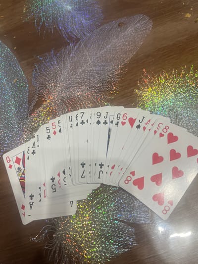 A fanned out deck of playing cards rests on a wooden surface decorated with iridescent, feather-like patterns. The cards, mostly white with red and black suits, display various ranks including Ace, 5, 6, 7, 8, 9, and Jack. Some cards are clearly visible, such as the Queen of Diamonds and an Ace of Clubs. The background shows a wooden table with holographic or glittery feather designs. The lighting suggests an indoor setting, possibly a home. There is no indication of location within the city of Rosenovo, Bulgaria.