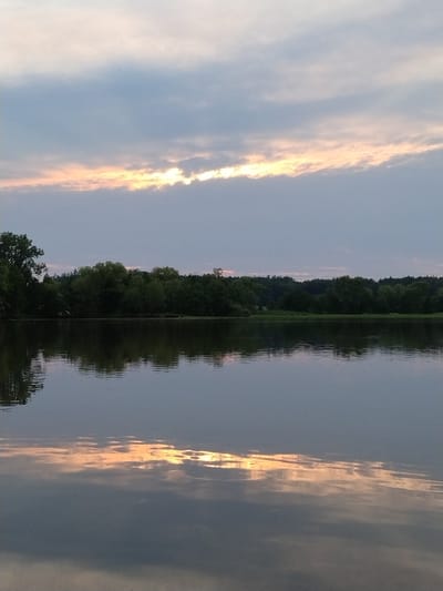 The image depicts a tranquil lake at dusk, reflecting the sky and surrounding trees. A line of dense trees borders the far side of the lake, with the sky above showing soft clouds tinged with the warm hues of a fading sunset. The water's surface is gently rippled, creating a mirror-like effect that distorts the reflection of the clouds and trees. The scene is set in Kestřany, Czechia, as indicated by the provided context. There are no people or discernible actions captured in the frame, rendering the scene a serene, natural landscape.