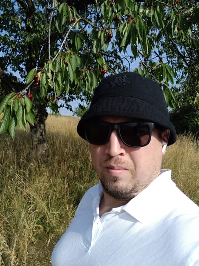 A man wearing a black bucket hat, sunglasses, and a white polo shirt takes a selfie in an outdoor setting. He has a light beard and is wearing white wireless earbuds. Behind him, a cherry tree with ripe red cherries and green leaves is visible against a bright blue sky. The ground is covered in tall, dry, golden-brown grass. The scene suggests a warm, sunny day in a rural or natural environment. The time of day appears to be daytime, likely late morning or afternoon, given the brightness and shadows. The location is identified as Šternov, Czechia. There is no visible text in the image.