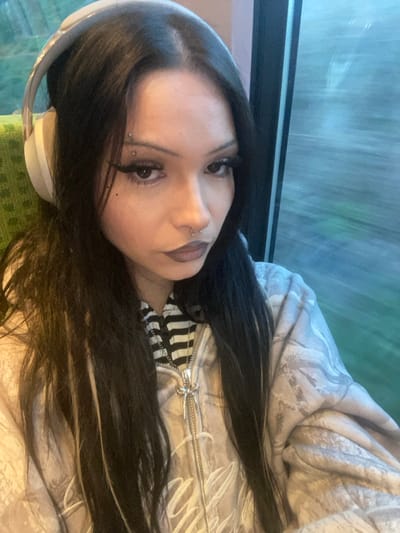 A young female is depicted in a close-up, selfie-style photograph taken inside what appears to be a moving train. She has long, dark hair with lighter streaks, centrally parted, and wears light-colored over-ear headphones. Her face features distinct winged eyeliner, prominent eyelashes, dark-toned lipstick, a silver septum piercing, and a horizontal piercing above her left eyebrow. Two small black adornments are visible below the outer corner of her left eye. She is dressed in a black and white striped undergarment beneath a light beige zippered hoodie. The hoodie features a patterned design and clearly displays the word "Enghien" on its front. A silver chain necklace with an elaborate pendant, including a small blue gem, is visible around her neck. The background through the train window exhibits horizontal motion blur, consistent with travel, and the interior visible behind her left shoulder shows green upholstered seating. Based on the "Enghien" text on her clothing and the train setting, the subject is located on a train associated with Enghien-les-Bains, France.