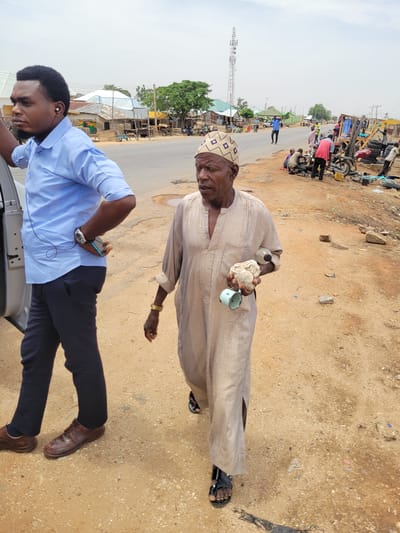 The image depicts two men in what appears to be Mara, Nigeria. An older man wearing a tan robe and patterned cap holds a white solid and a small blue cup. He also wears sandals. Beside him, a younger man in a blue shirt and black pants is leaning on what seems to be a vehicle. He has an earpiece and is looking at his phone. The ground is sandy and dusty. In the background, there is a road with people and vehicles, including motorcycles. A communication tower is visible in the distance, along with buildings and vegetation.