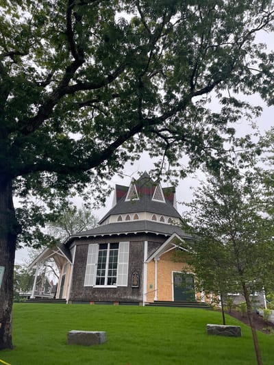 The image shows the Federated Church of Martha's Vineyard in Oak Bluffs, Massachusetts, United States of America. The church is a historic octagonal building with a distinctive red roof and white trim. The building is surrounded by lush green grass and mature trees. The sky is overcast. The church is the focal point of the image, with the trees framing it on either side. A sign is visible on the side of the church with text that reads "1870 CHAPEL". There are two large stones in the foreground, and a smaller tree is visible to the right of the church. The Federated Church is a prominent landmark in Oak Bluffs, known for its unique architecture and historical significance.