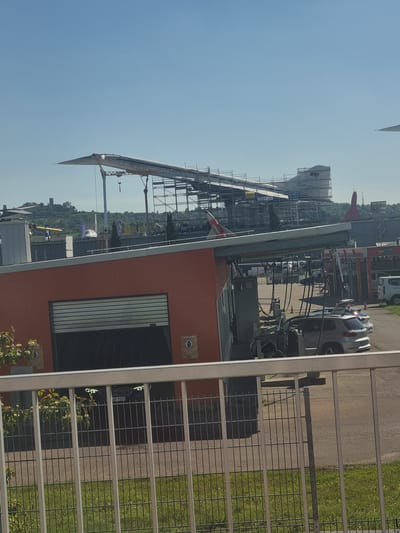 The image shows a partial view of the Technik Museum Sinsheim in Sinsheim, Germany. In the foreground, a fence obscures a part of a building painted in orange. The building has a large window with vertical blinds. The background contains elements of the museum's exhibits, including the Concorde airplane mounted atop a structure, a large tank with "CCCP-77108" printed on it, and other displays. Cars and structures are also visible in the distance, and the sky is blue.