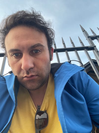 A low-angle, close-up, eye-level selfie captures a person from the chest up, facing the camera. The individual has dark, curly hair, blue eyes, and is wearing a bright blue jacket over a yellow shirt. Sunglasses are resting on the yellow shirt, with a reflection of buildings in the lenses. The background consists of a black metal fence with pointed finials, and a cloudy sky. The location is the city of Armagh, United Kingdom.