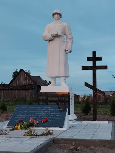 This is a medium shot photograph of a war memorial with a soldier statue and a wooden Orthodox cross. The statue, made of white material, depicts a soldier in a helmet and a long coat, holding binoculars and a book or map. The cross, made of dark wood, is an eight-pointed cross, common in Eastern Orthodox tradition, and is positioned to the right of the statue.
In front of the statue is a dark, angled plaque with names and dates inscribed, suggesting it's a tombstone or memorial listing those who died. Below the plaque, a bouquet of flowers, some red and some yellow, lies on a grey tiled surface. The overall scene appears to be a cemetery or a memorial site.
Behind the memorial, there's a rustic wooden building with a steeply pitched roof, characteristic of rural architecture. The sky is overcast with a pale blue hue, suggesting either early morning or late evening. The setting appears to be outdoors in a rural or semi-rural area, possibly in Belarus given the context clue. The atmosphere is somber and reflective, fitting for a war memorial. The visible text includes "Здесь похоронен" (Here lies buried) and a poem that speaks of immortal hymns and the glory of fallen heroes.