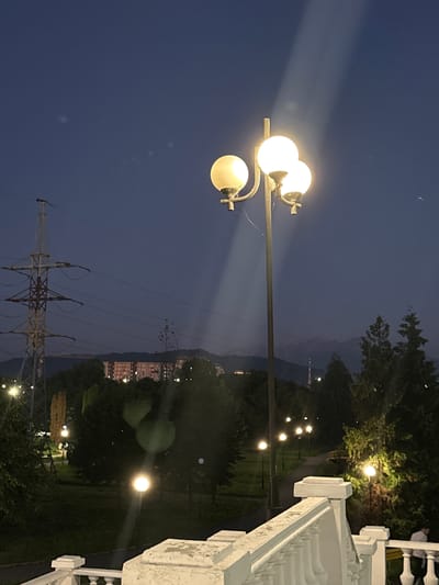 The image depicts a view of Arkabash 1-Y, Russia, at dusk. In the foreground, a white ornate railing and steps lead the eye into the scene. A multi-globe streetlight illuminates the immediate area with a warm glow. Beyond the railing, a park landscape unfolds with trees and lampposts lining a winding path. In the middle ground, a tall power pylon stands prominently against the twilight sky, with a residential building visible behind it. Distant mountains are silhouetted against the horizon, their peaks partially obscured by the fading light. The sky is a deep blue, with a few faint white specks, possibly distant lights or atmospheric phenomena. The overall atmosphere is serene and tranquil.