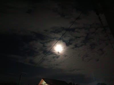 The image shows a nighttime sky with a bright moon partially obscured by clouds. The moon is positioned in the upper center of the frame, emitting a strong glow that illuminates the surrounding clouds. Two dark power lines traverse the sky diagonally, crossing in front of the moon. Below the sky, the silhouette of a building with a dark roof and a light-colored facade is visible at the bottom center. The lower part of the image is darker, with hints of vegetation or other structures on either side of the building. The scene appears to be outdoors, possibly in a residential area, given the presence of the building and power lines. The photograph was taken in the city of San Pascual, Philippines, suggesting a tropical night sky. There are no people or discernible actions taking place in the image.