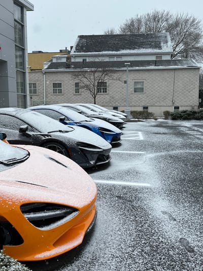 The image captures an outdoor scene in Greenwich, United States, during a light snowfall. A row of high-end sports cars, including one prominent orange vehicle in the foreground, are parked in an asphalt lot and lightly dusted with snow. The cars behind the orange one are primarily dark-colored (blue, gray, black), also showing a layer of fresh snow. White parking lines are visible on the asphalt, partially obscured by the snow, and some footprints can be seen on the ground.
In the background, two buildings are prominent. The larger building features a light-colored, possibly shingled or scaled, exterior and a dark, snow-covered roof. It has multiple rectangular windows. To its left, another building with a more modern, glass facade is partially visible. Bare trees, indicative of winter, stand between and behind the buildings, their branches outlined against the overcast sky. Snowflakes are still visible in the air, suggesting ongoing or very recent snowfall. The overall lighting is even, typical of an overcast daytime. No discernible text is visible in the image.