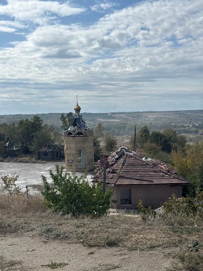 This image depicts a damaged landscape in Izium, Ukraine, specifically around the Holy Spring of the Pishchanska Icon of the Mother of God. The scene is observed from an elevated, unpaved vantage point, characterized by dry, sparse vegetation in the immediate foreground.
In the midground, a light-colored, octagonal brick or stone chapel structure with a blue and gold onion dome stands prominently. The dome exhibits significant damage, with numerous blue tiles missing and exposed structural elements. An Orthodox cross is visible atop the dome. The chapel's arched openings are empty, lacking windows or doors, and some graffiti-like markings are present on its lower walls, including partially visible Cyrillic text and a blue emblem. To the right of the chapel, a single-story brick building with a gabled, red tiled roof is severely compromised; multiple tiles are missing, exposing the underlying wooden rafters, and sections of the roof appear collapsed. An open doorway or window on its façade reveals interior debris. Further to the left and slightly behind the chapel, a smaller, dilapidated wooden hut with a partially collapsed roof is visible. The ground immediately surrounding these structures is light-colored, appearing to be bare earth, rubble, or dried mud. Interspersed among the buildings and in the foreground are various bushes and trees, some green and others exhibiting autumnal brown foliage.
The background consists of extensive rolling hills covered in dense green and brown woodland, stretching to the horizon under a sky with scattered cumulus clouds and patches of clear blue. No people are visible in the image.