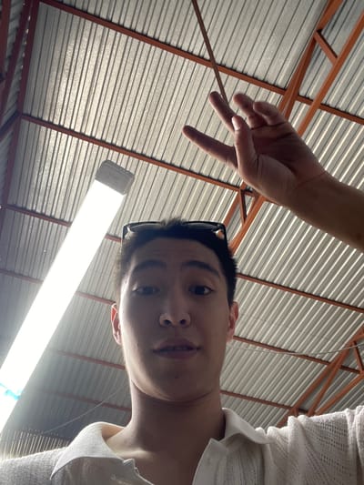 A low-angle, medium shot captures a young Asian male from the chest up, looking directly at the camera. He has short black hair and is wearing a white collared shirt. Sunglasses are perched on top of his head. His left hand is raised in a V-sign gesture, with his fingers extended towards the ceiling. His right hand is not fully visible.
The background consists of a corrugated metal roof supported by orange metal beams. A long, bright fluorescent light fixture runs diagonally across the upper left of the frame. The overall environment appears to be indoors, possibly a workshop, warehouse, or covered outdoor space. The lighting is strong from the overhead fixture. The image was taken in Кенен Әзірбаев ауылы, Kazakhstan.