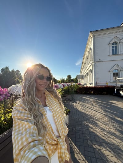 An outdoor scene in Dumino, Russia, depicts a woman in the foreground taking a selfie during late afternoon or early evening. She possesses long, wavy blonde hair with integrated braids, wears dark oval sunglasses, and is smiling faintly while dressed in a yellow and white gingham patterned shirt over a white top. The sun is positioned behind and to her left, creating a prominent lens flare and illuminating her hair.
To the left of the woman, green trees and bushes are visible, along with arrangements of white and pink large-blossomed flowers, likely hydrangeas, displayed in planters along a dark metal railing. A section of a dark wooden bench is partially visible in the lower-left corner.
On the right side of the image, a large, multi-story white building with classical architectural elements, including arched windows, decorative cornices, and pilasters, dominates the background. A dark sloped roof is visible at the top. A paved pathway of grey rectangular pavers extends from the mid-ground to the lower right, marked by long, distinct shadows cast from the building and unseen elements, indicative of the low sun angle. Dark reddish-brown bushes line the base of the building, where a security camera is mounted. A white golf cart or utility vehicle is parked near the building's facade on the far right. A coiled black cable or hose lies on the pathway in the mid-ground, and a partial yellow warning sign is visible near the building.