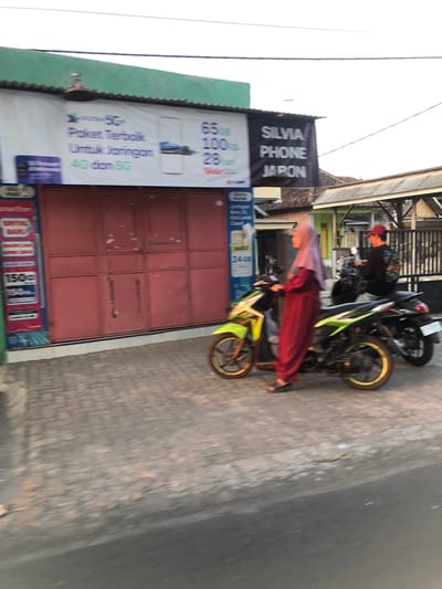 In Sentul, Indonesia, a street scene unfolds with a focus on a retail establishment and its immediate surroundings. The building's facade features a large, red, corrugated metal door and a prominent banner advertising mobile phone plans, including "5G Paket Terbaik Untuk Jaringan 4G dan 5G." Adjacent to this, another banner proclaims "SILVIA PHONE JARON." On the left side of the building, a vertical sign displays pricing information with denominations such as "150" and "750." In the foreground, two individuals are on motorbikes, suggesting movement and transit. One person, wearing a purple hijab and a red dress, is on a green and black motorbike. Behind them, a second person wearing a red cap and a dark patterned shirt is on a black motorbike. The ground is paved with cobblestones, and the overall lighting suggests it is either early morning or late afternoon.