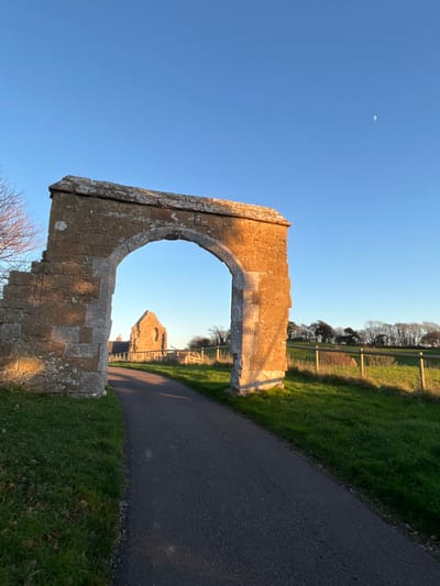 The image displays a clear daytime scene at the ruins of Abbotsbury Abbey, located in Abbotsbury, Dorset, United Kingdom. The foreground is dominated by a substantial stone archway, likely part of the Great Gatehouse, constructed from large, light-colored blocks with visible weathering and partial deterioration on its left flank. Direct, golden-hour sunlight illuminates the archway's right side and interior curve, casting deep shadows on the left.
A dark, asphalt pathway runs from the bottom center, passing directly through the archway and extending into the mid-ground. Beyond the arch, further along the path, stands a distinct, dilapidated stone structure, consistent with a remaining section of the abbey church. Green grass borders the path on both sides. To the right of the path and beyond the archway, a wooden post-and-wire fence separates the path from a verdant green field. In the background, a line of bare deciduous trees stretches across a slight rise. The upper left corner shows a portion of a tree with bare branches.
The sky is uniformly clear and blue, with a slender crescent moon visible in the upper right quadrant. No individuals, animals, or active events are present in the depicted scene.